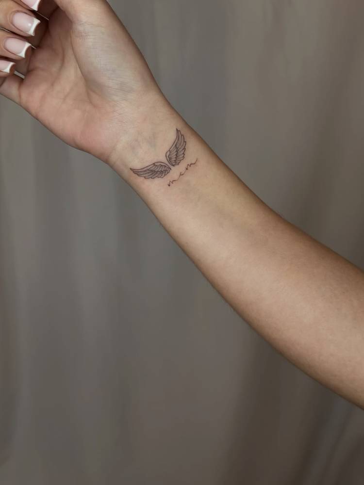 Fine line style angel wings and initials tattoo located on the wrist.