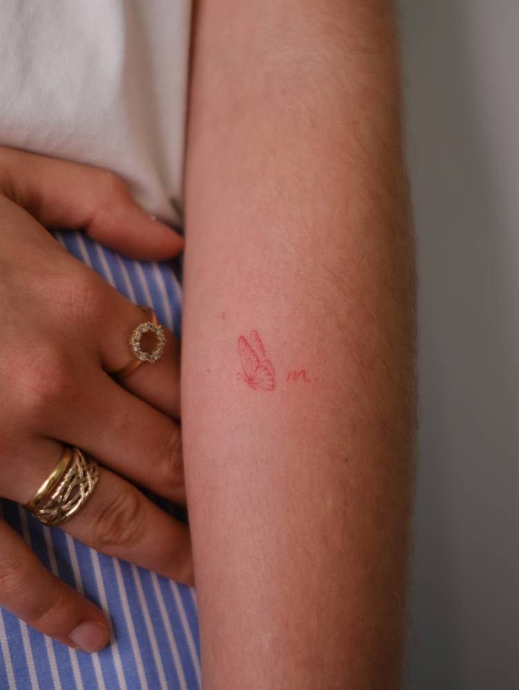 Red ink butterfly and letter "M" tattoo located on the inner forearm.