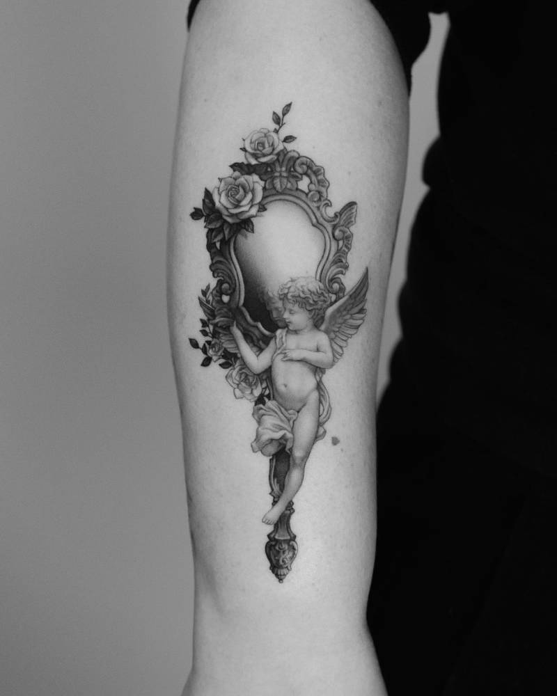 Classical style cherub and mirror tattoo located on the forearm.