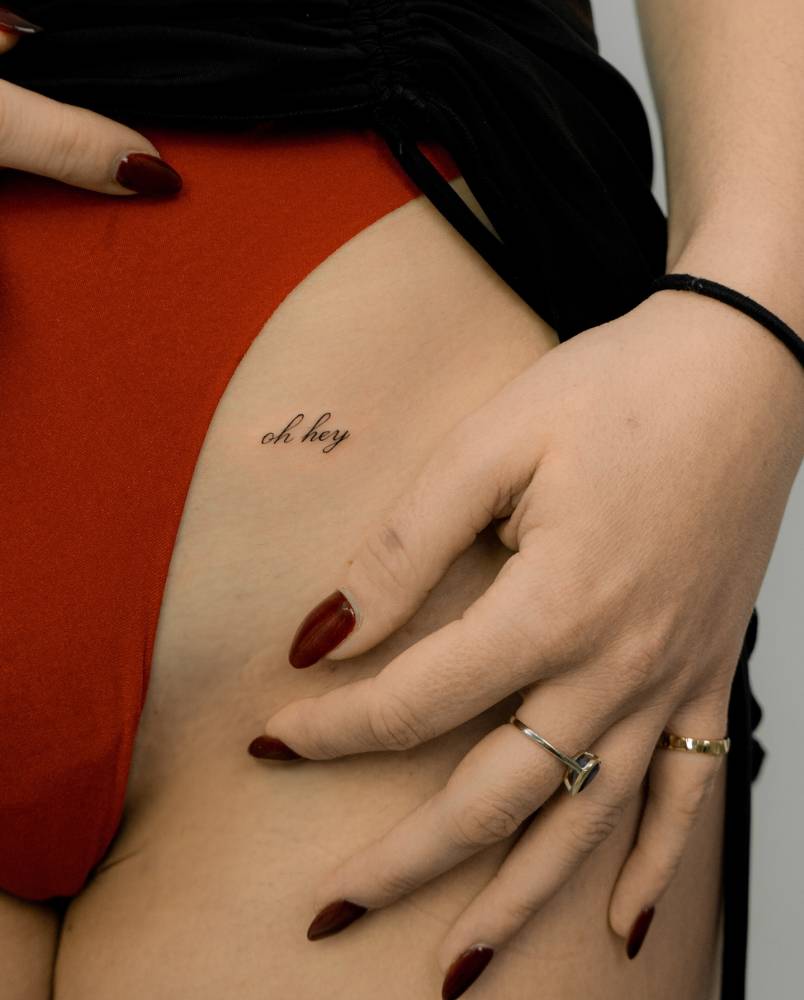 Script font "Oh hey" lettering tattoo located on the pelvis.
