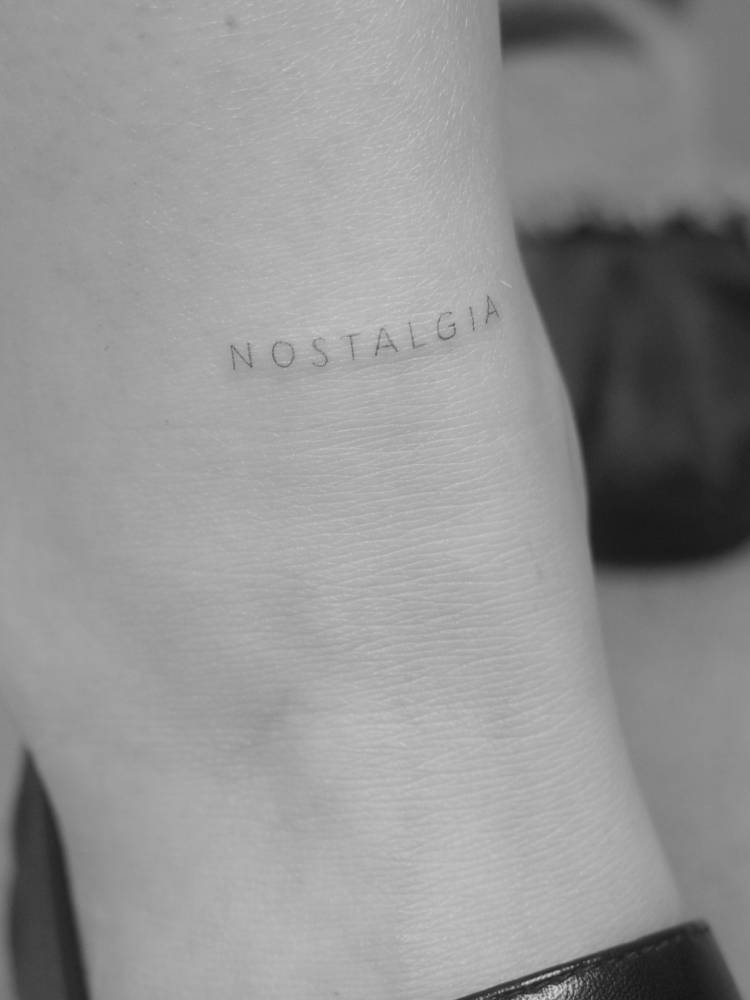 Word "nostalgia" lettering tattoo located on the ankle.