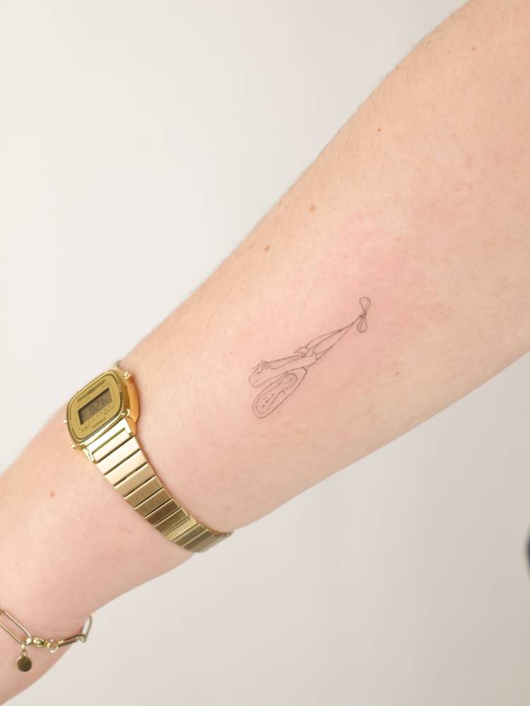 Ballet shoes tattooed on the forearm, done in fine line style.