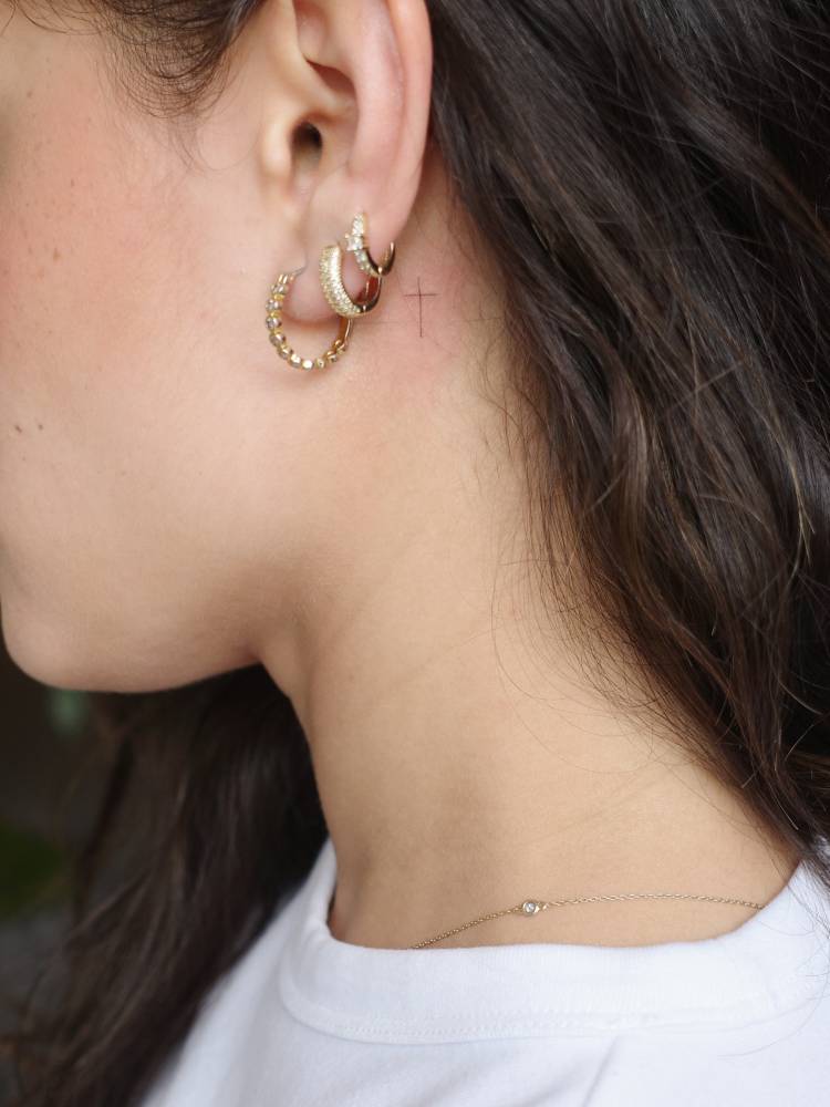 Tiny minimalist cross tattoo placed behind the ear.