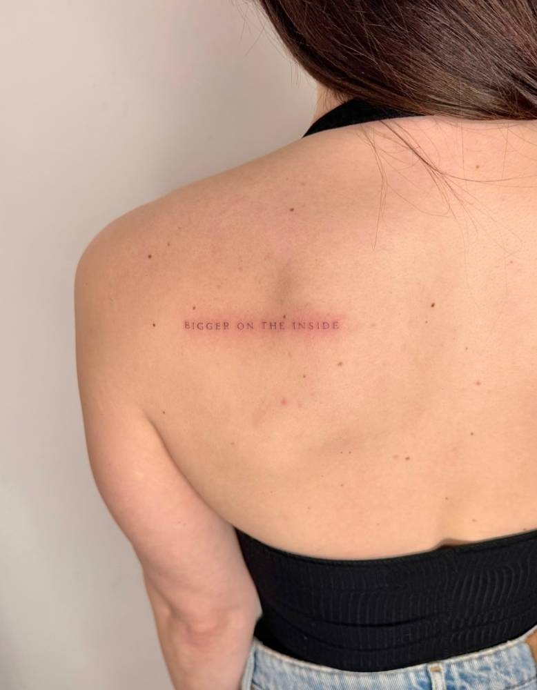 "Bigger on the inside" lettering tattoo located on the shoulder blade.