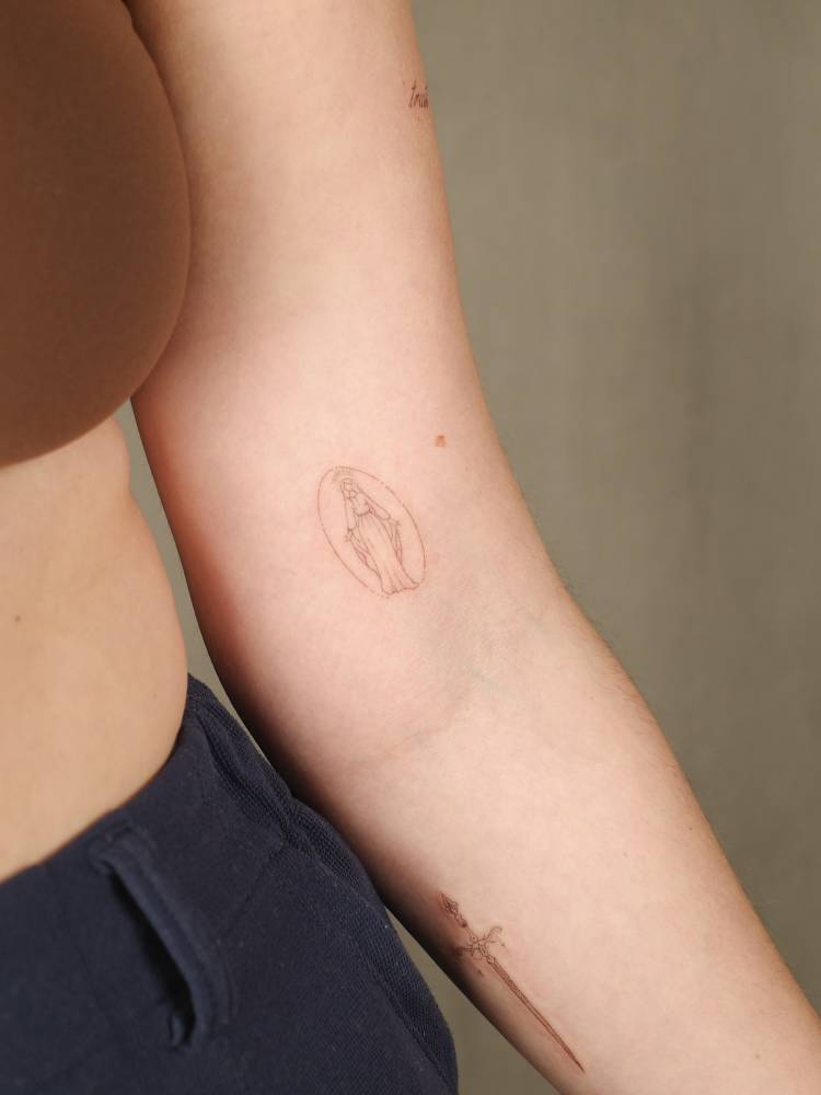 Tiny Virgin Mary tattoo in fine line style located on the inner arm.