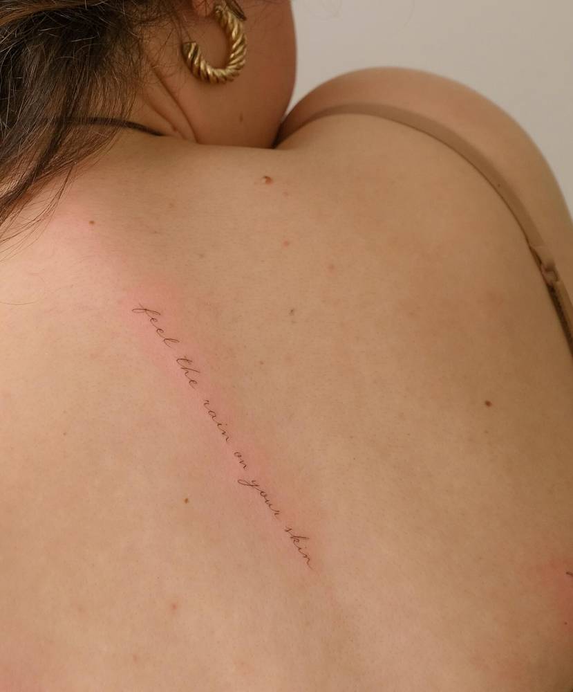 "Feel the rain on your skin" lettering tattoo located on the upper back.