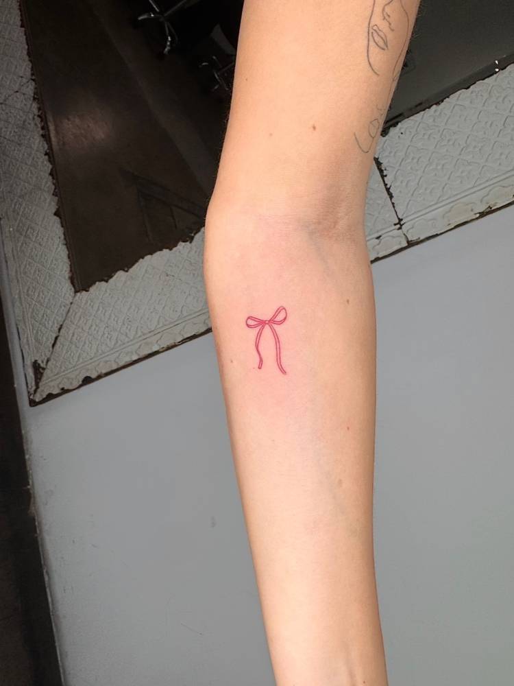 Red bow tattoo in fine line style placed on the inner forearm.
