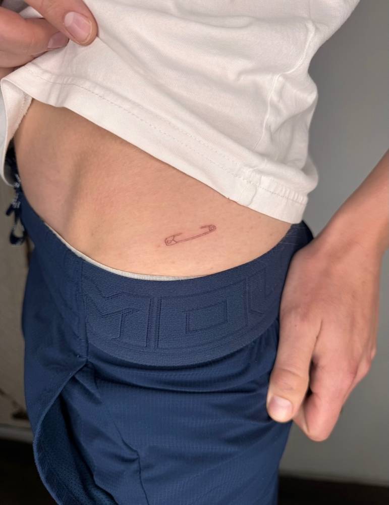 Fine line style safety pin tattoo located on the hip.