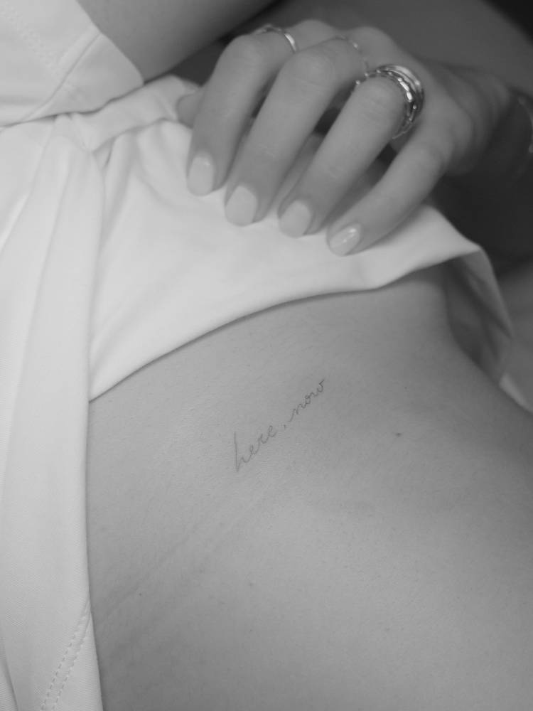 Handwritten font "here now" lettering tattoo done on the rib.