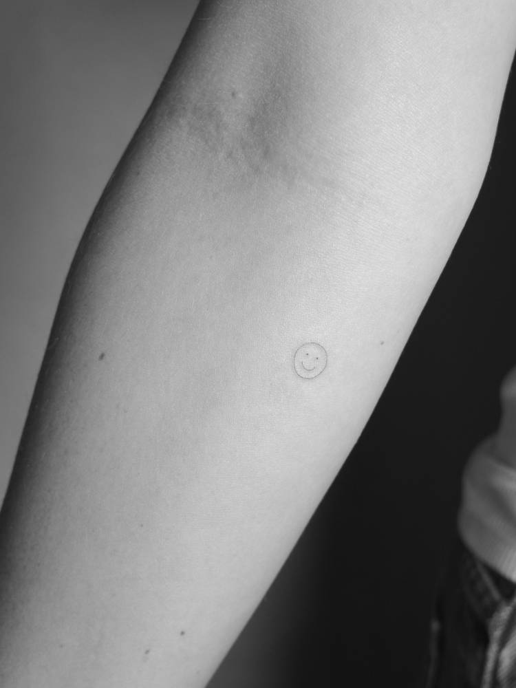 Tiny smiley face tattoo in minimalist style located on the inner forearm.