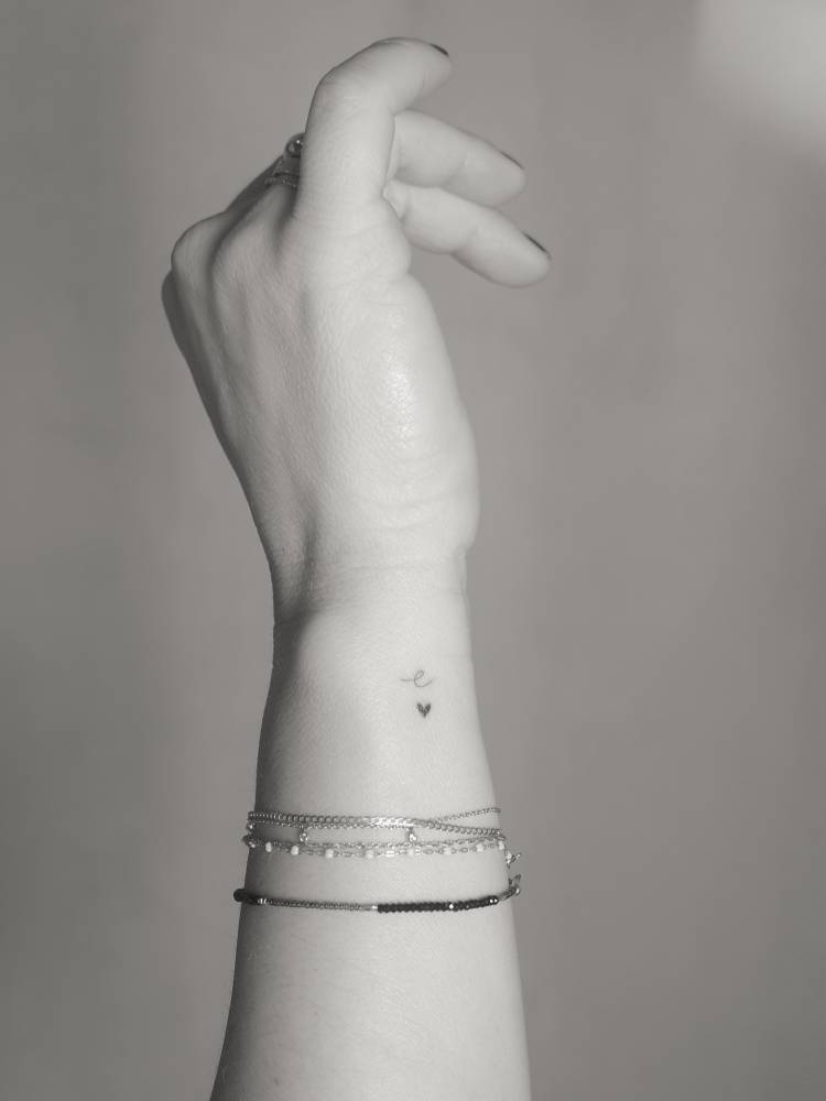 Tiny letter "e" and heart tattoo in minimalist style placed on the wrist.