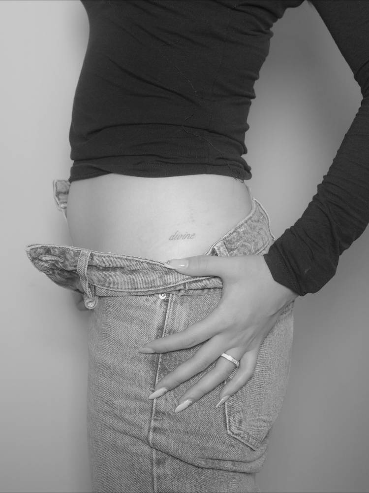 Script font word "divine" tattooed on the hip.