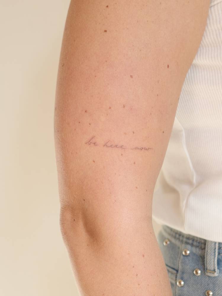 Handwritten font "be here now" lettering tattoo located on the upper arm.