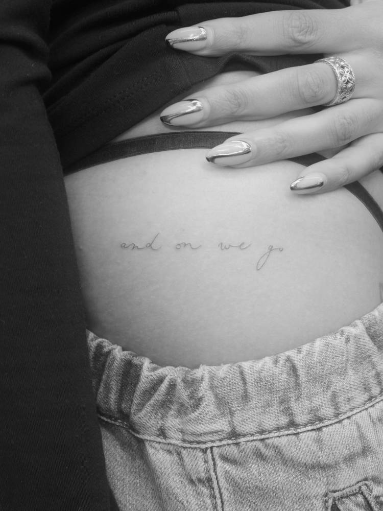 "And on we go" in handwritten font tattooed on the hip.