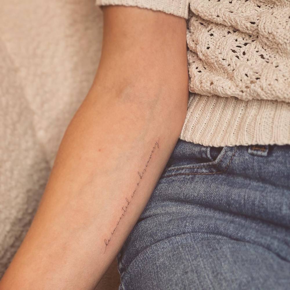 "Be invested in a discovery" lettering tattoo located on the inner forearm.
