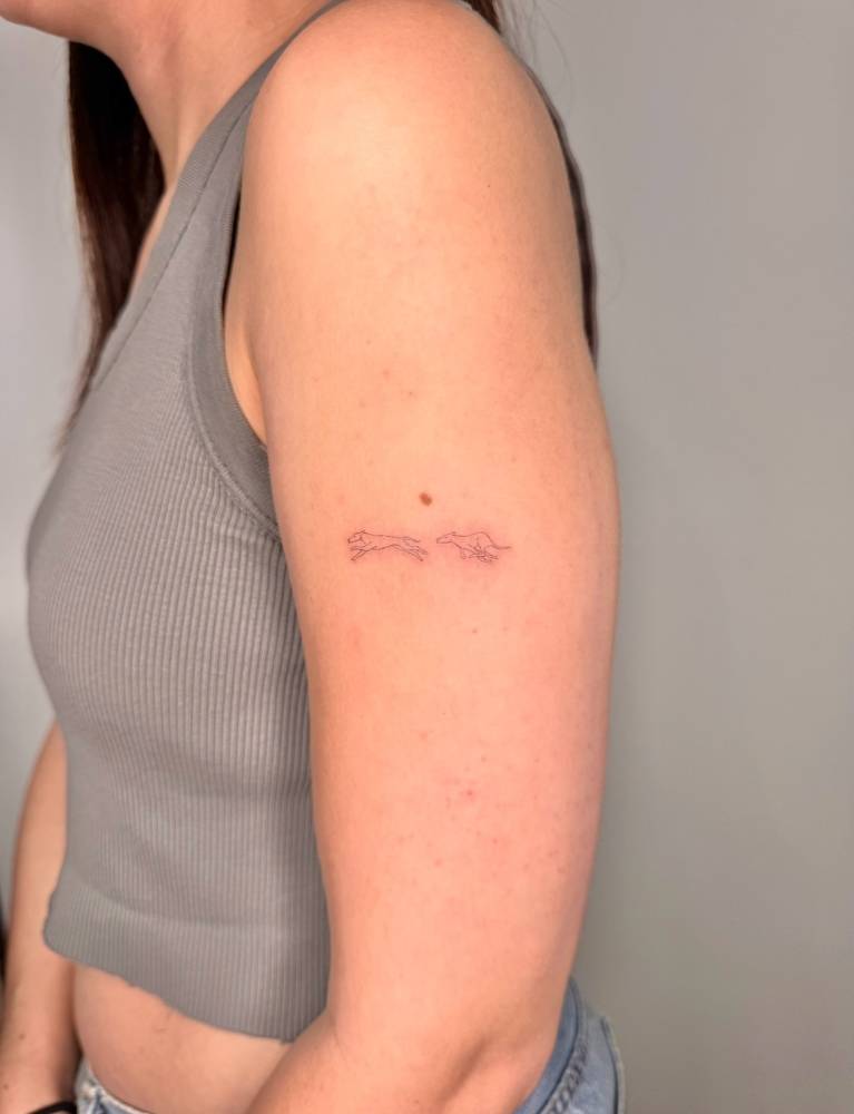 Little running greyhounds tattoo placed on the upper arm, fine line style.