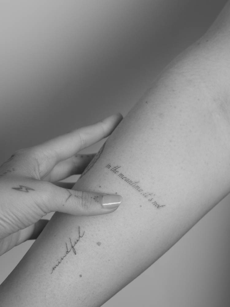 "In the meantime, it's not" lettering tattoo in script font.
