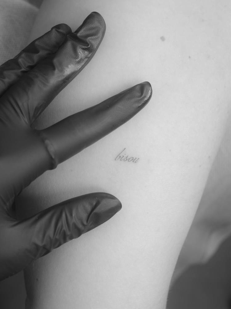 Script font word "bisou" tattooed on the upper arm.