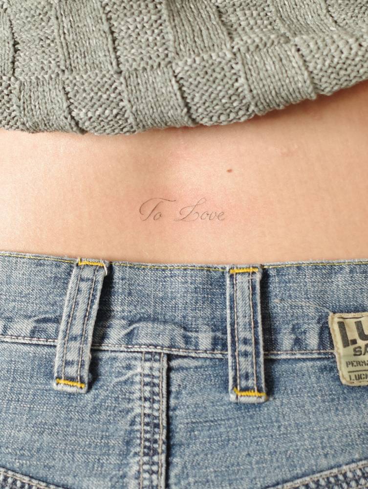 "To love" lettering tattoo located on the lower back.