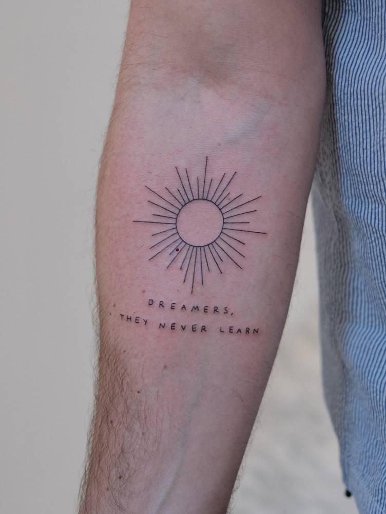 "Dreamers, they never learn" and sun tattooed on the inner forearm.