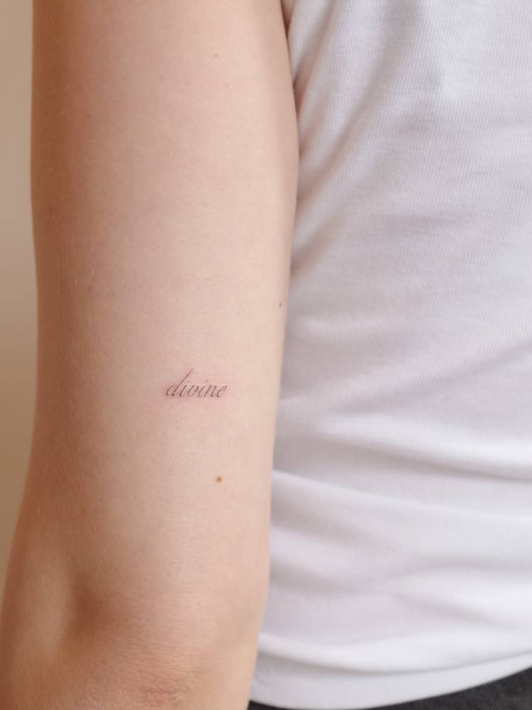 Script font word "divine" tattooed on the tricep.