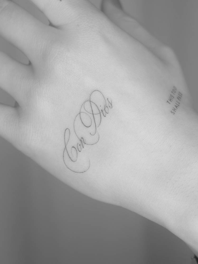 Script font "Con Dios" lettering tattoo located on the hand.