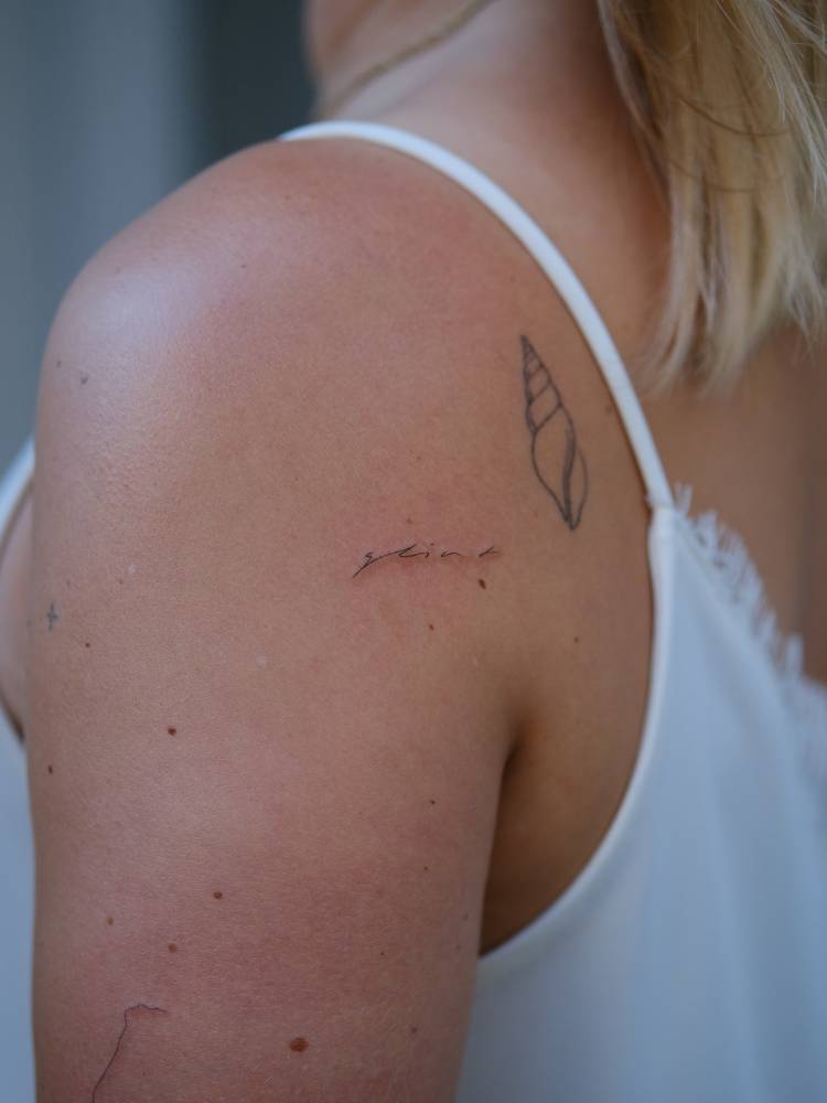 "Glint" lettering tattoo in handwritten font located on the shoulder.