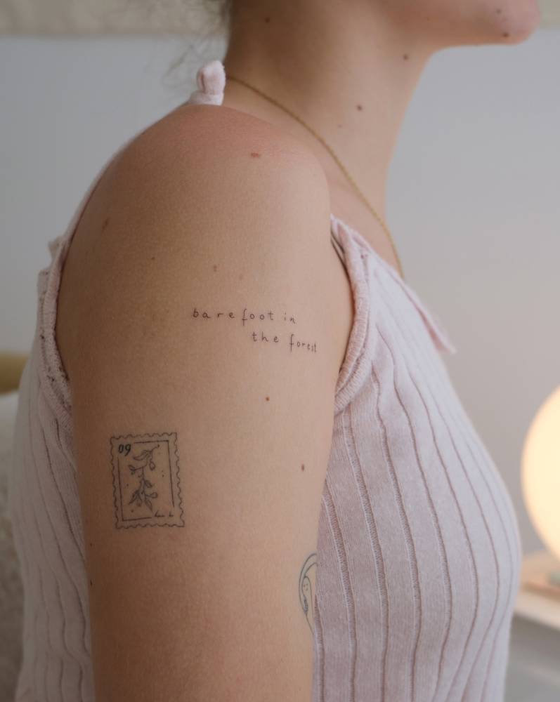 "Barefoot in the forest" lettering tattoo located on the upper arm.