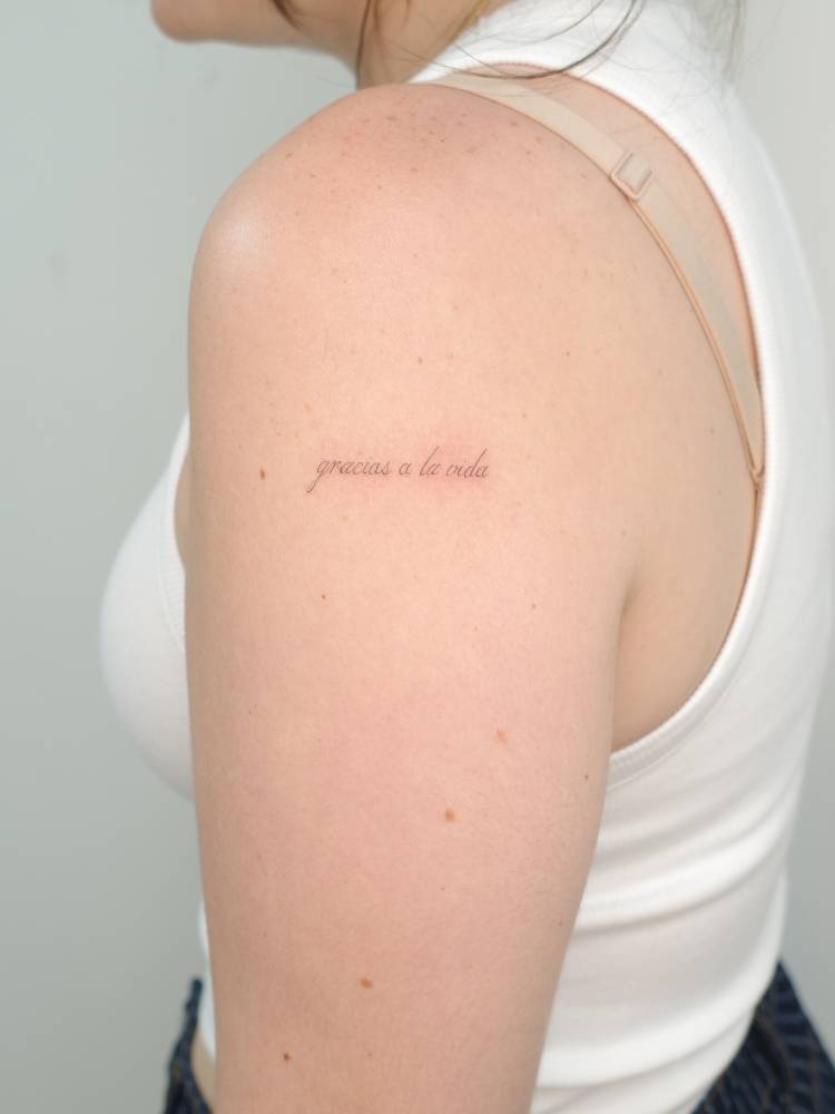 "Gracias a la vida" lettering tattoo placed on the upper arm.