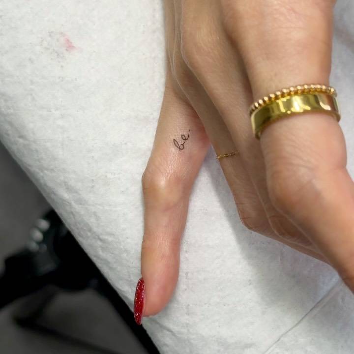 Handwritten font word "be" tattooed on the finger.