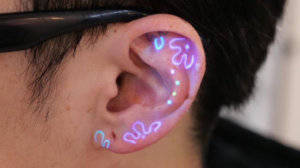 UV ink flowers and dots tattooed on the ear, minimalistic style.