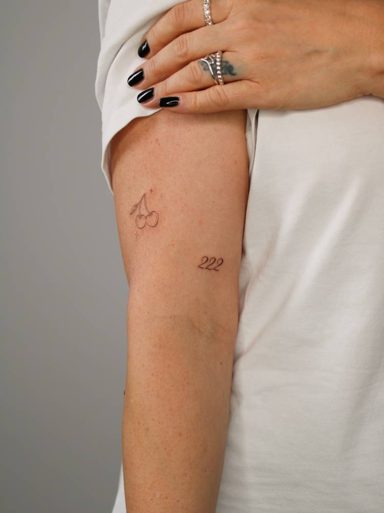 Fine line style cherries and angel number "222" tattooed on the upper arm.
