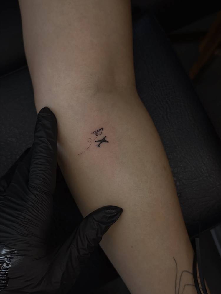 Paper plane and airplane shadow tattoo located on the inner forearm.