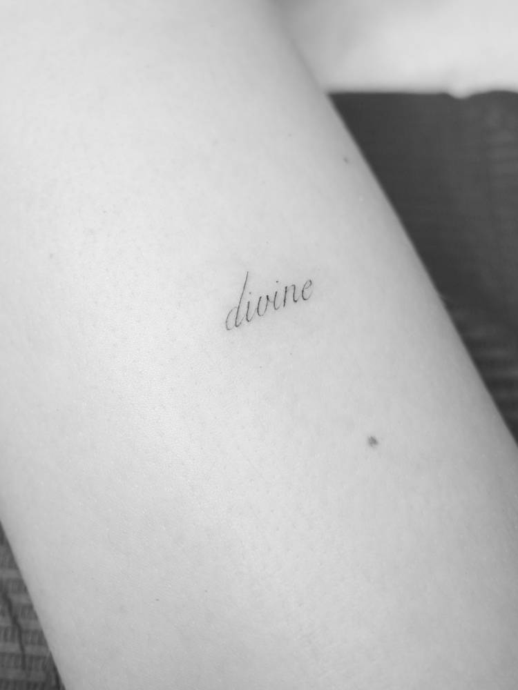 Script font word "divine" tattooed on the upper arm.