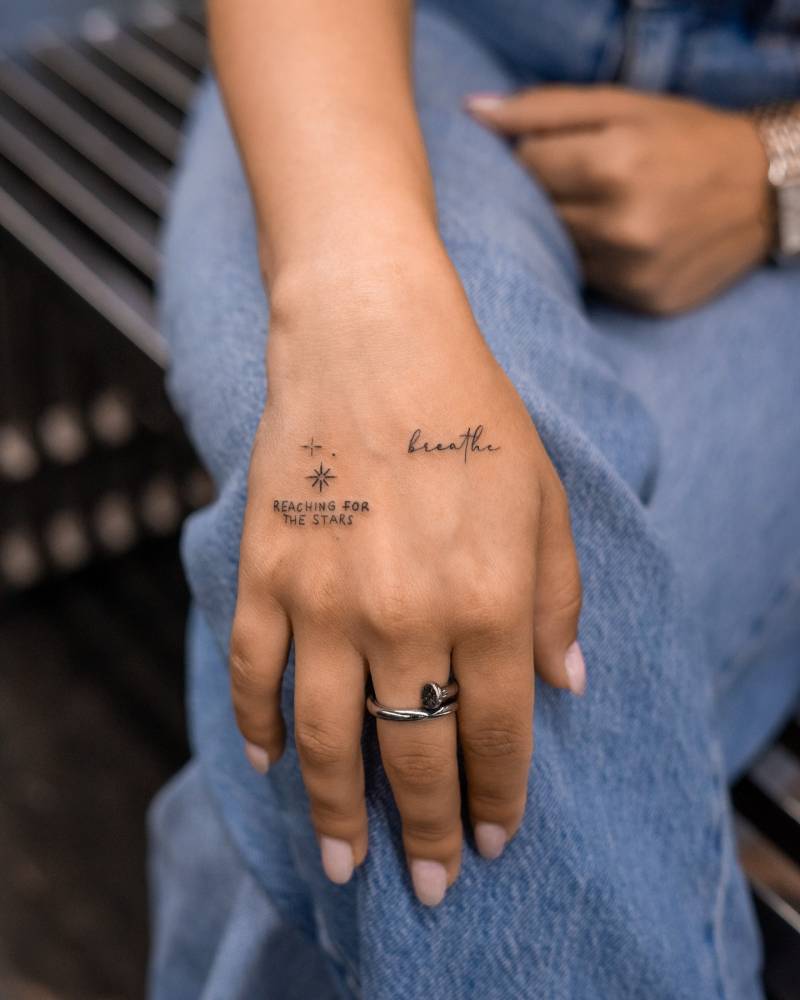 "Reaching for the stars" and "breathe" lettering tattoos located on the hand.