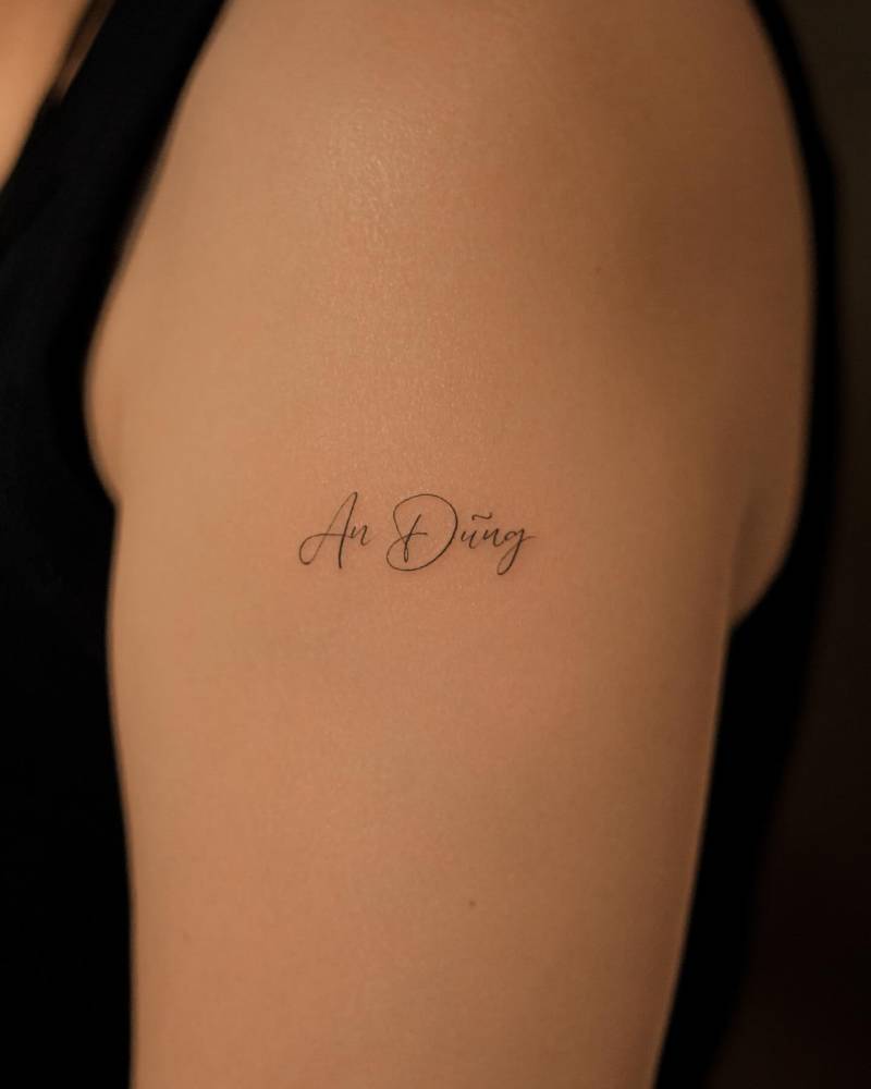 "An Dũng" lettering tattoo in serif font located on the upper arm.