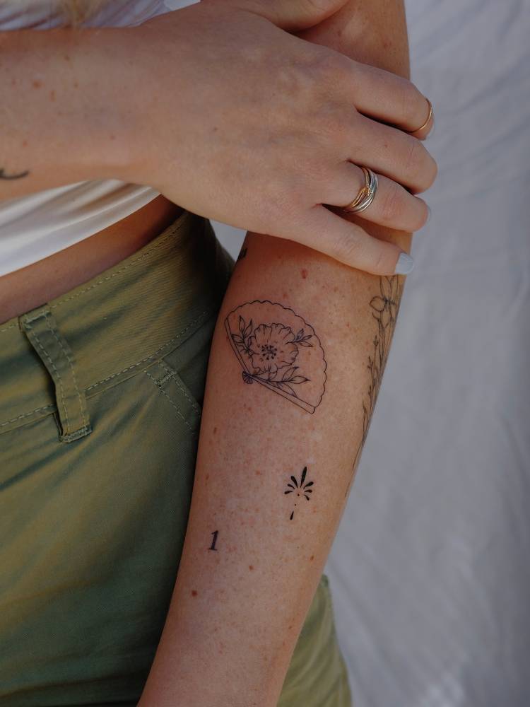Hand fan, ornament and number "1" tattoos located on the forearm.