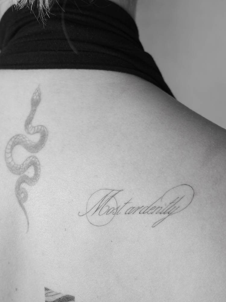 "Most ardently" lettering tattoo in script font located on the shoulder blade.