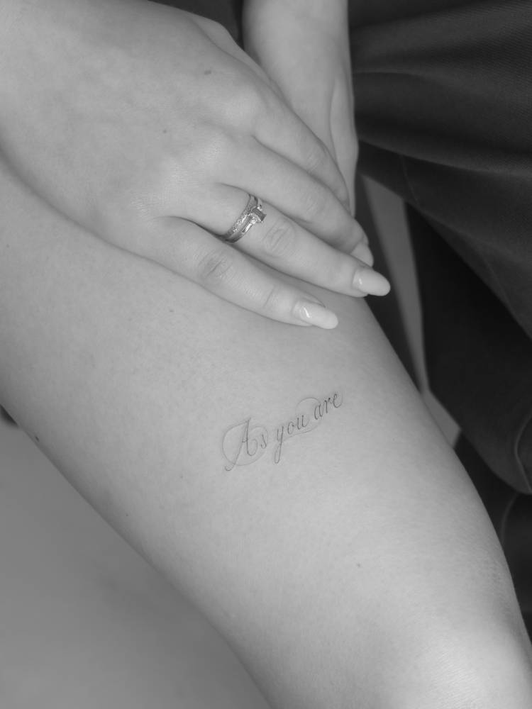 Script font "as you are" lettering tattoo located on the thigh.