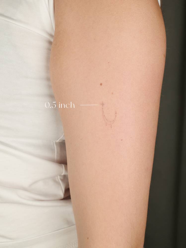 Tiny ornamental moon tattooed on the upper arm, fine line style.