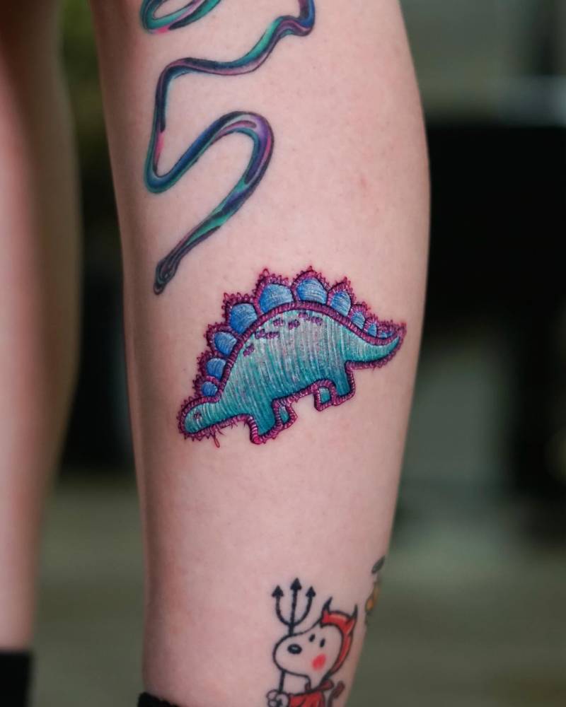 Stegosaurus patch tattoo in embroidery style placed on the shin.