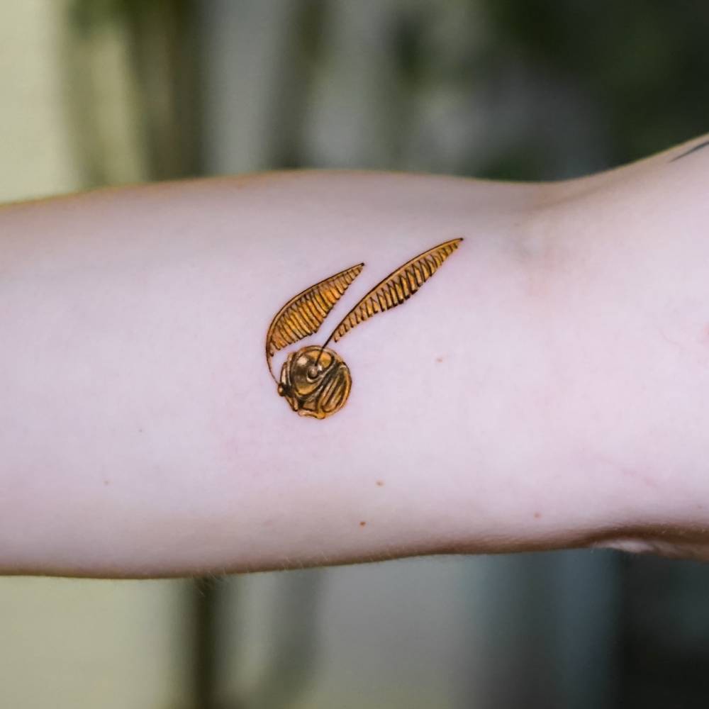 Golden Snitch tattoo in micro-realistic style placed on the inner forearm.