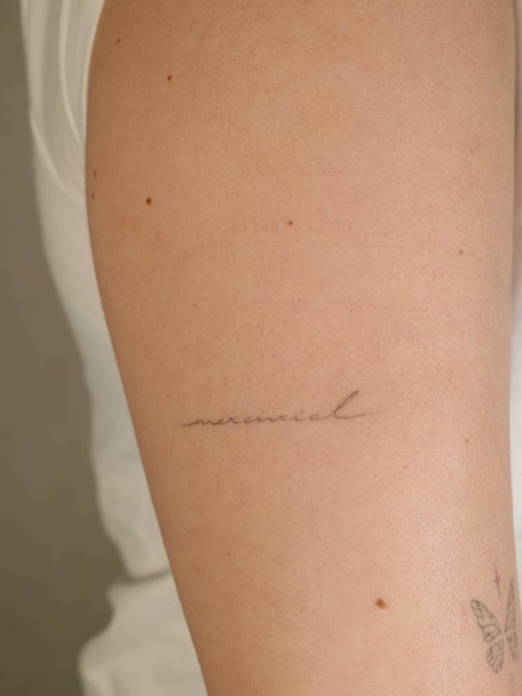 Handwritten font word "mercurial" tattooed on the upper arm.