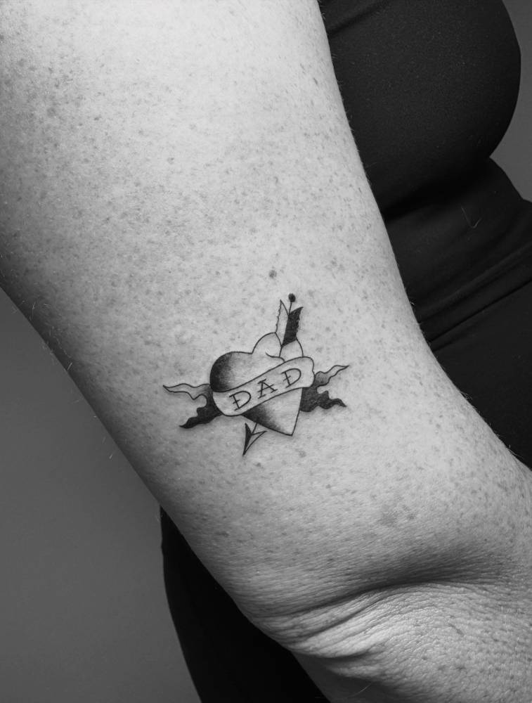 Heart and dagger with "dad" banner tattooed on the upper arm.