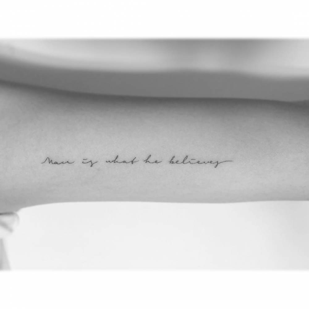 "Man is what he believes" lettering tattoo handwritten on the inner arm.