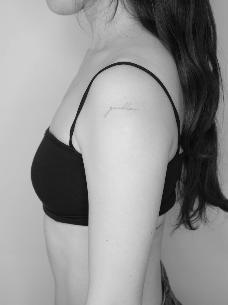 Handwritten font word "gentle" tattoo located on the shoulder.