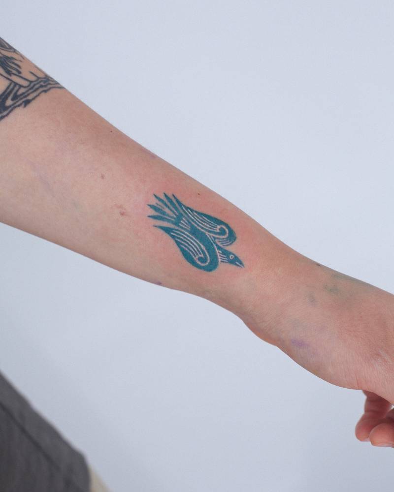 Stylized bird tattoo located on the wrist, done in blue ink.