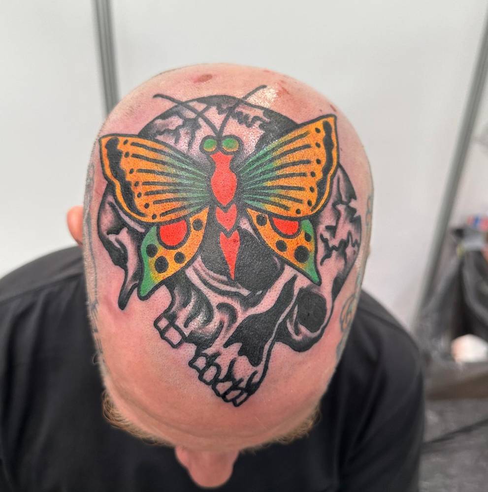 Skull and butterfly tattoo placed on the top of the head, traditional style.