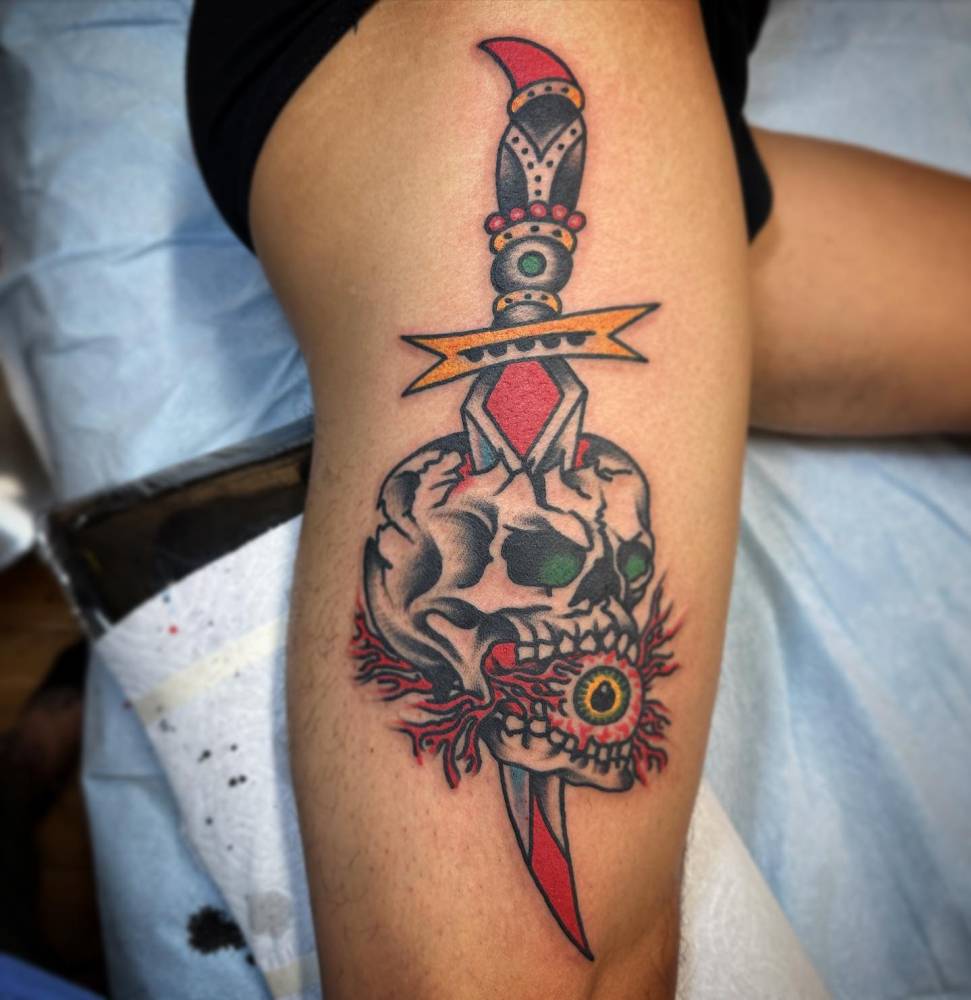 Skull and dagger tattoo located on the thigh, traditional style.