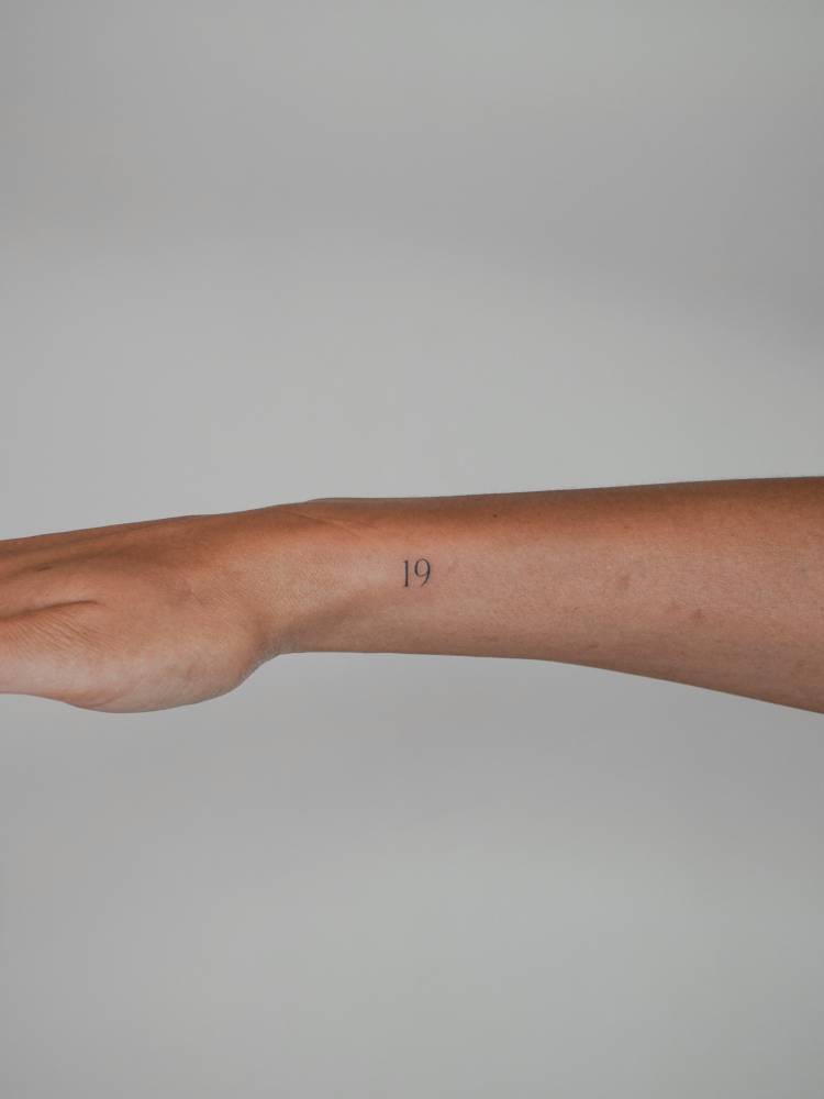 Tiny number "19" tattoo in serif font located on the wrist.